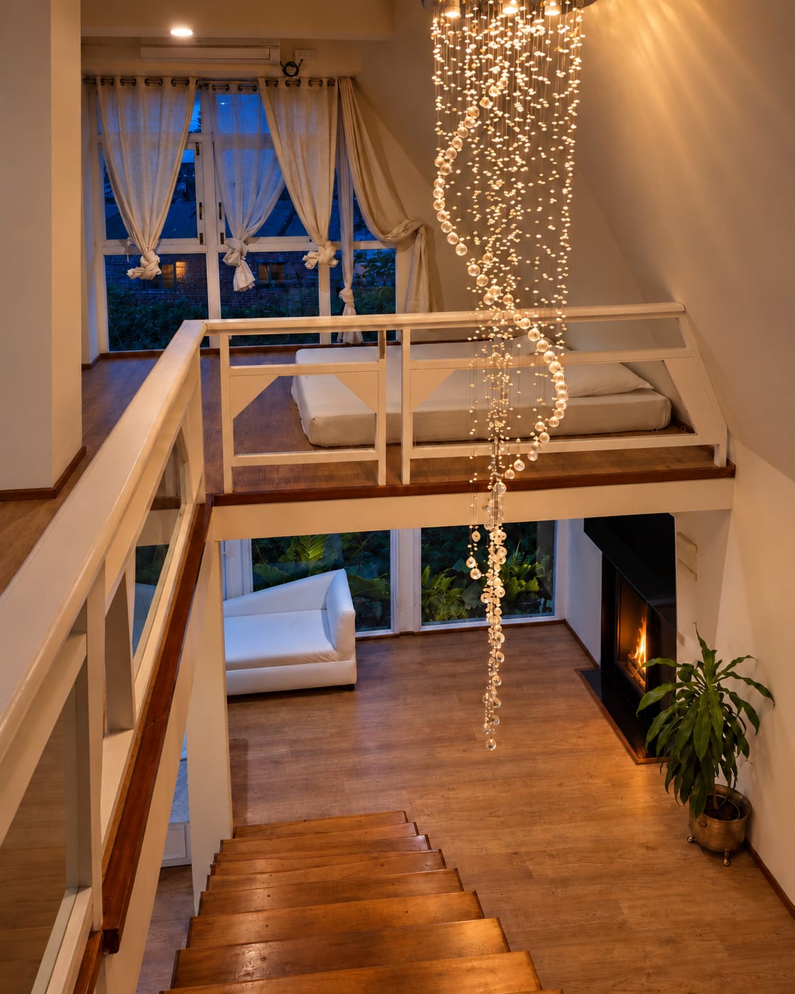 Looking down the homestay's timber stairs past the loft rail and a crystal chandelier, to the lounge and the fireplace below